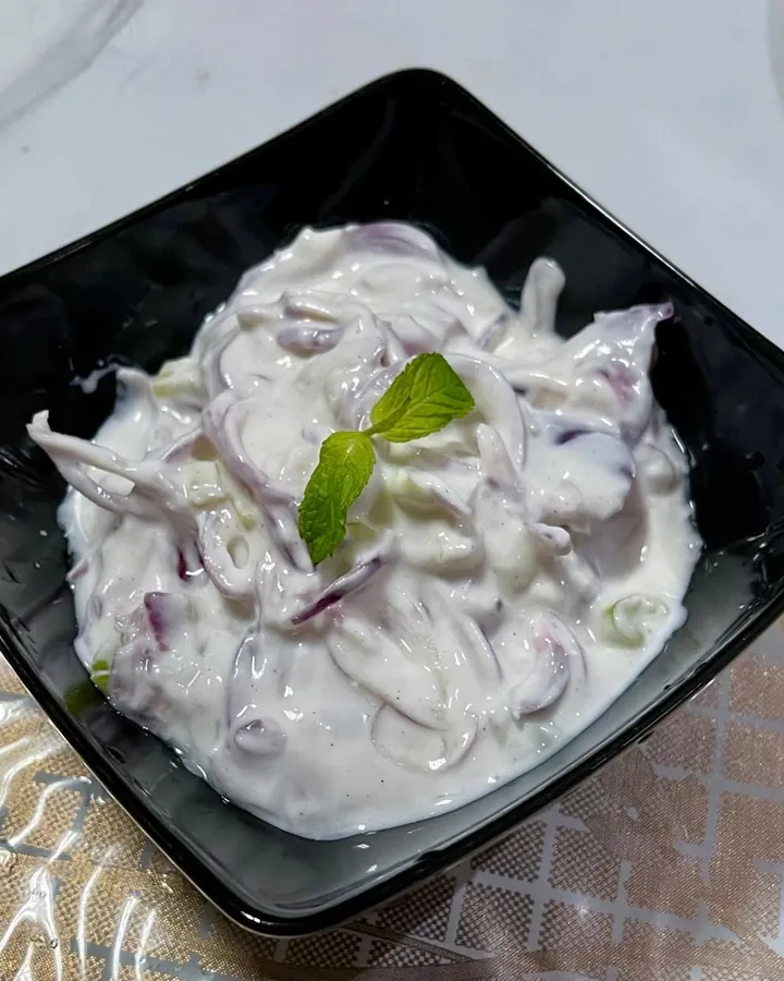 Creamy yogurt salad with thinly sliced red onion in a square black bowl, garnished with a sprig of mint