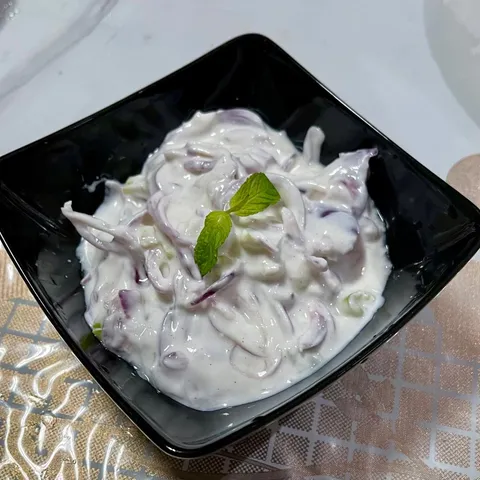Creamy yogurt salad with thinly sliced red onion in a square black bowl, garnished with a sprig of mint