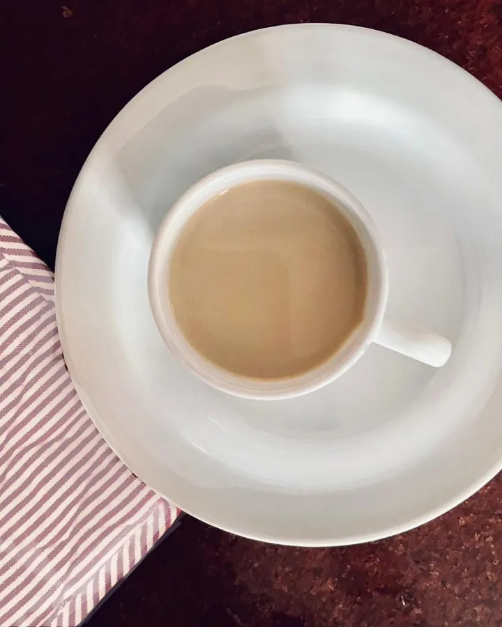 A white cup of creamy coffee milk tea on a white saucer beside a red-and-white striped napkin