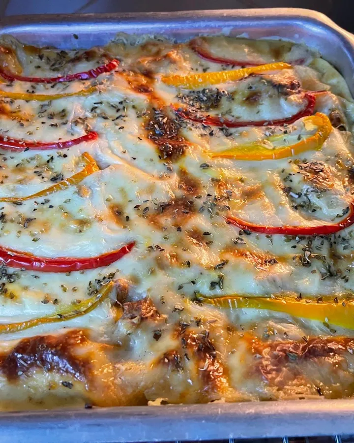 Baked lasagna in a metal tray, golden with browned spots on top and decorated with strips of red and yellow bell pepper and oregano