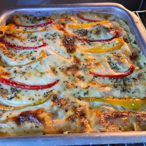 Baked lasagna in a metal tray, golden with browned spots on top and decorated with strips of red and yellow bell pepper and oregano