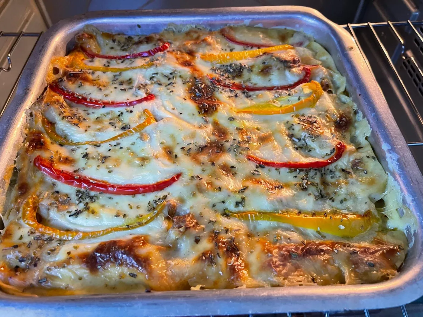 Baked lasagna in a metal tray, golden with browned spots on top and decorated with strips of red and yellow bell pepper and oregano