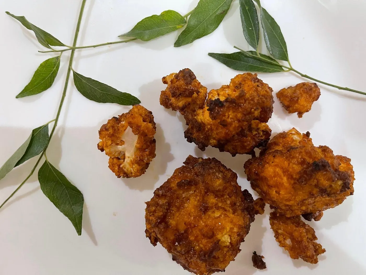 Crispy golden air-fried buffalo cauliflower bites on a white plate with sprigs of curry leaves