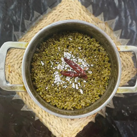 A pot of boiled green gram topped with scraped coconut and dried red chilies, on a woven mat