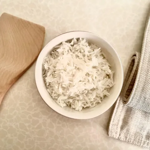 A white bowl of fluffy cooked white rice next to a wooden spatula and a linen napkin
