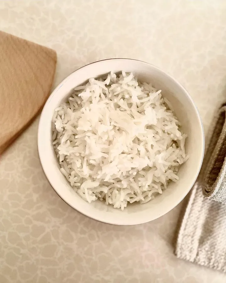A white bowl of fluffy cooked white rice next to a wooden spatula and a linen napkin