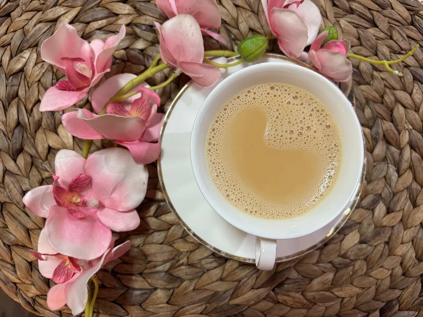 A cup of creamy milk tea on a white saucer, with pink orchids on a woven placemat