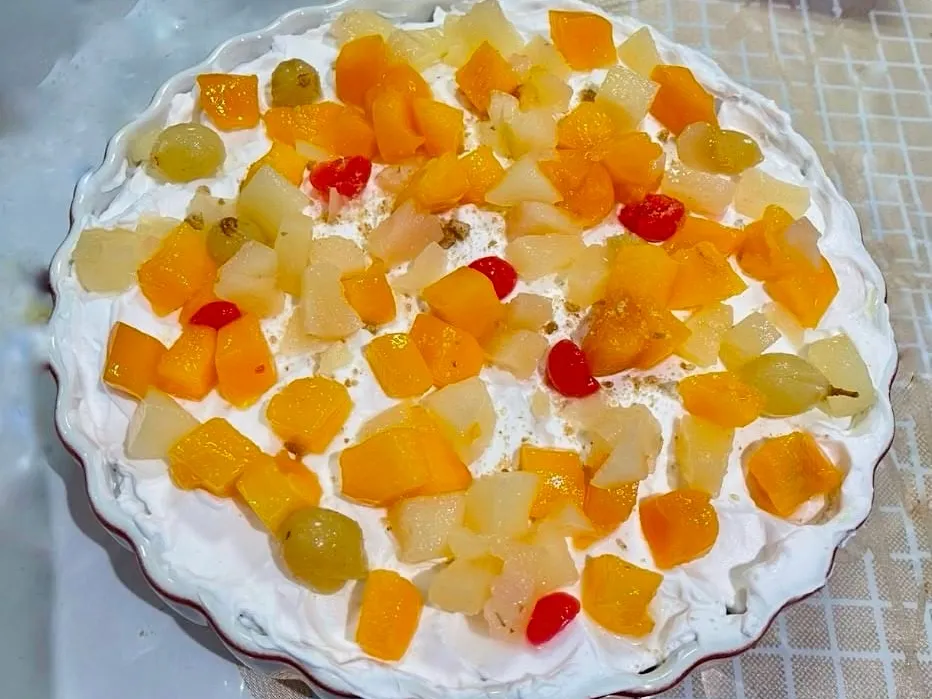 Fruit custard trifle in a white fluted dish, topped with whipped cream and colorful mixed fruit